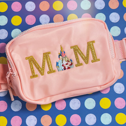 A pink fanny pack with the word 'MOM' embroidered in gold, featuring a castle and additional decorative elements.