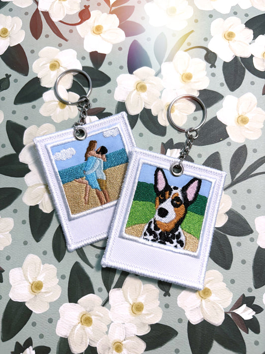 Two embroidered square keychains with silver key rings, displayed on a floral background. One keychain features an embroidered beach scene with two people, and the other features a black-and-white dog portrait.