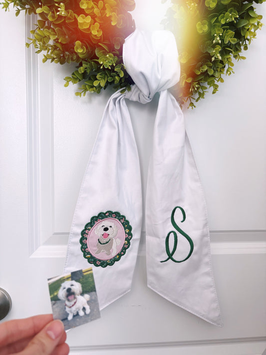 Wreath on a door that has a white wreath sash tied on it. Sash has embroidery of a dog on one side and “S” on the other.