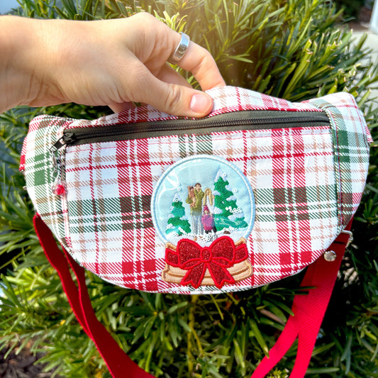 Green tree background with a red and green plaid crossbody in focus with an embroidered snow globe.