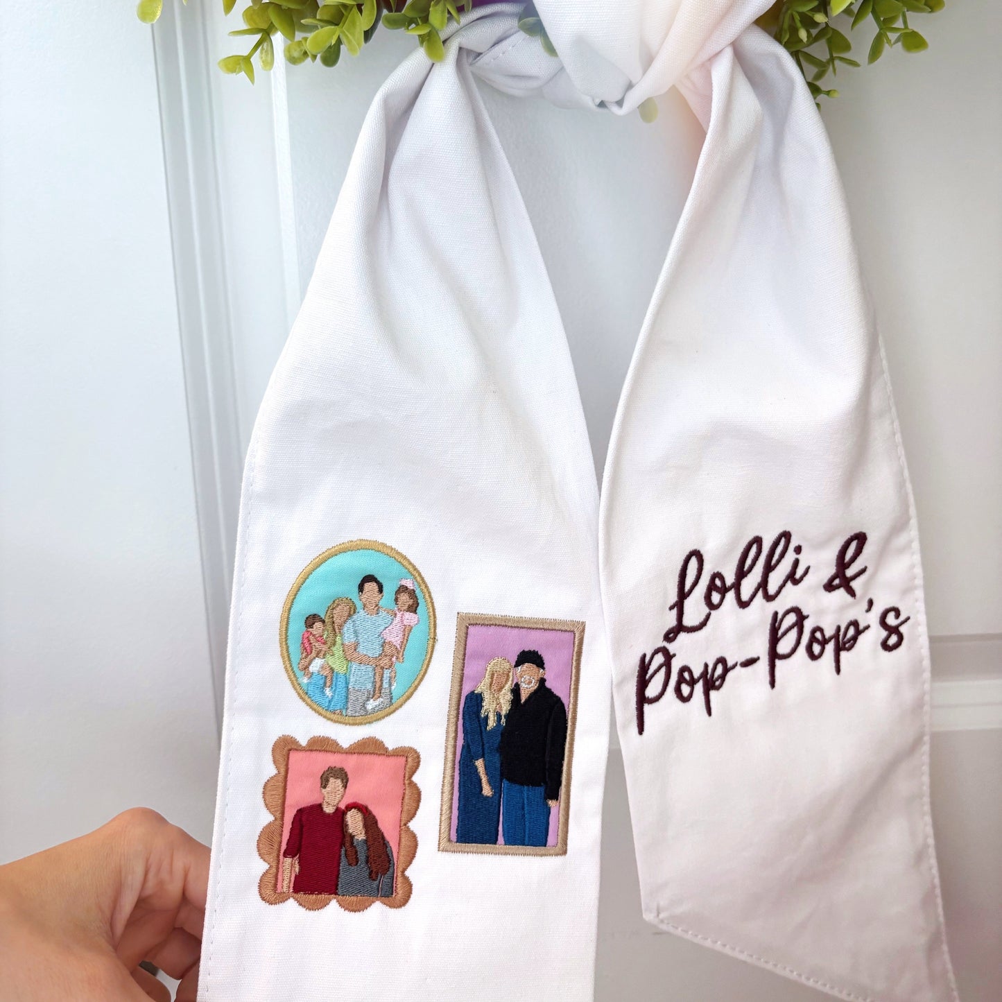 White scarf with embroidered designs and text held by a hand, with greenery in the background.