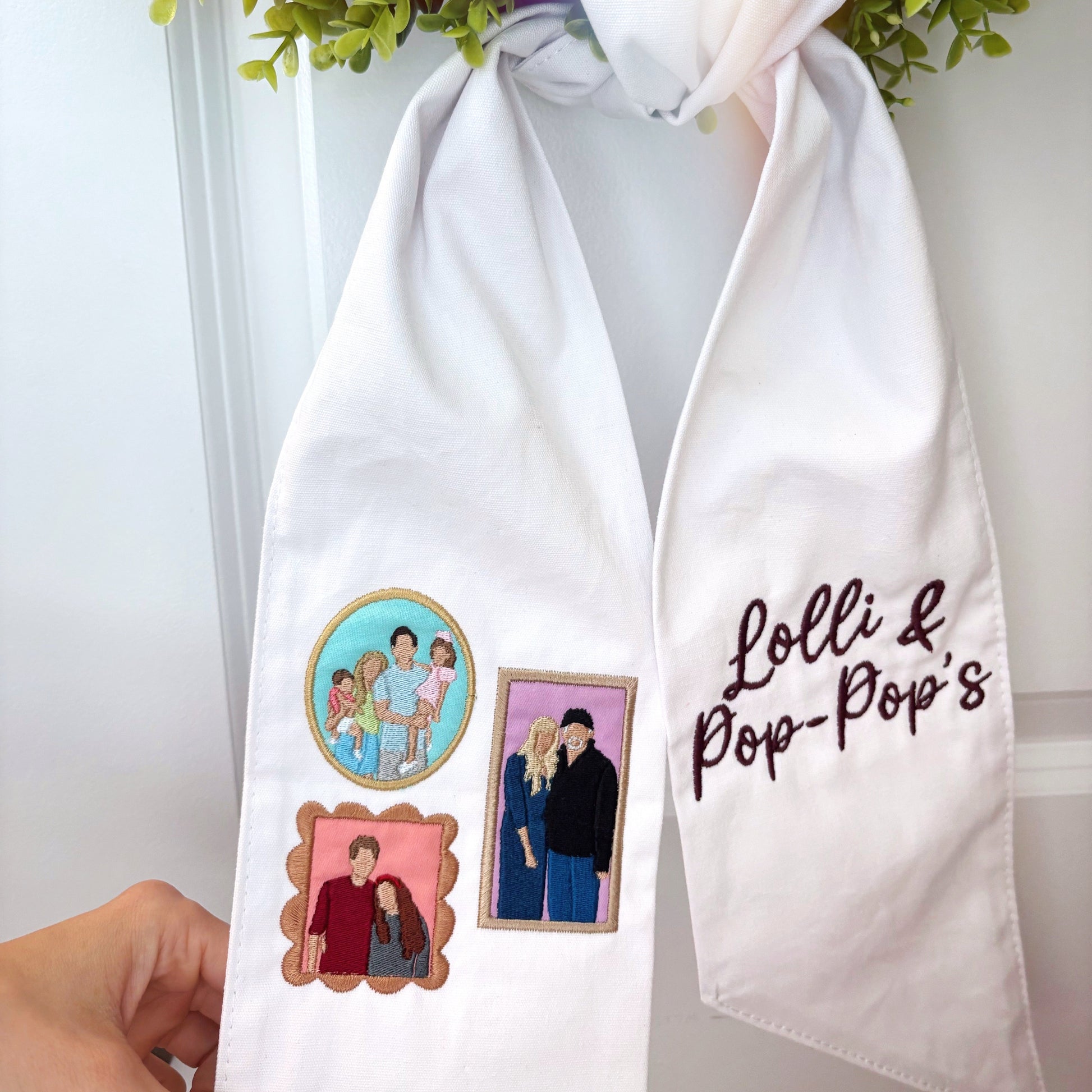 White scarf with embroidered designs and text held by a hand, with greenery in the background.