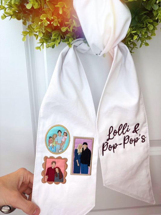 White scarf with embroidered designs and text held by a hand, with greenery in the background.