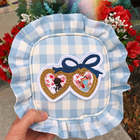 Blue gingham bag with a gold locket patch with two portraits and an embroidered blue bow. 