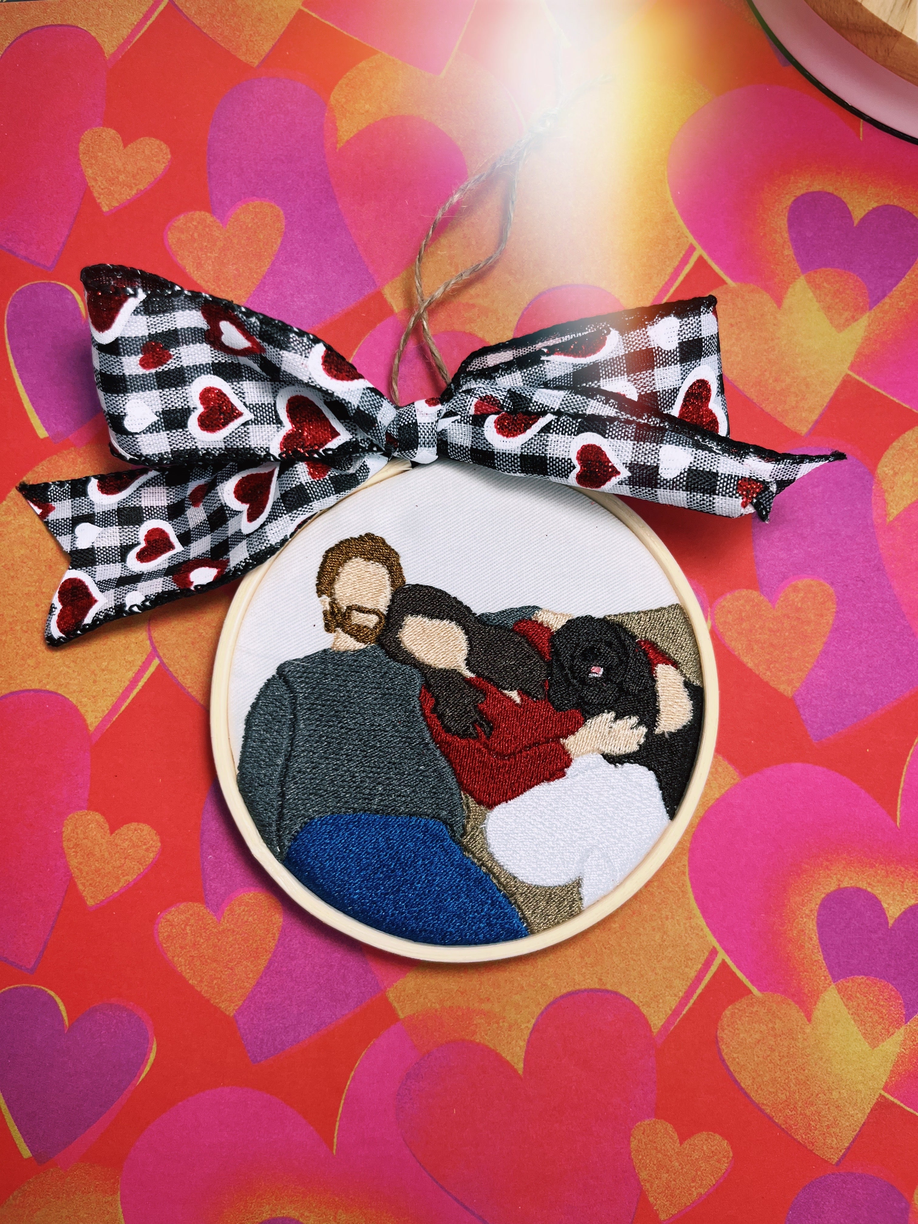 A custom embroidered ornament featuring an embroidered picture of two people with a dog, framed with a black and white checkered ribbon against a red and orange heart background.