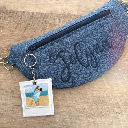 A personalized blue quilted fanny pack with a floral pattern, featuring the name 'Jolynn' embroidered in dark blue on the front, a top zipper, and a silver keyring with a custom embroidered photo patch.