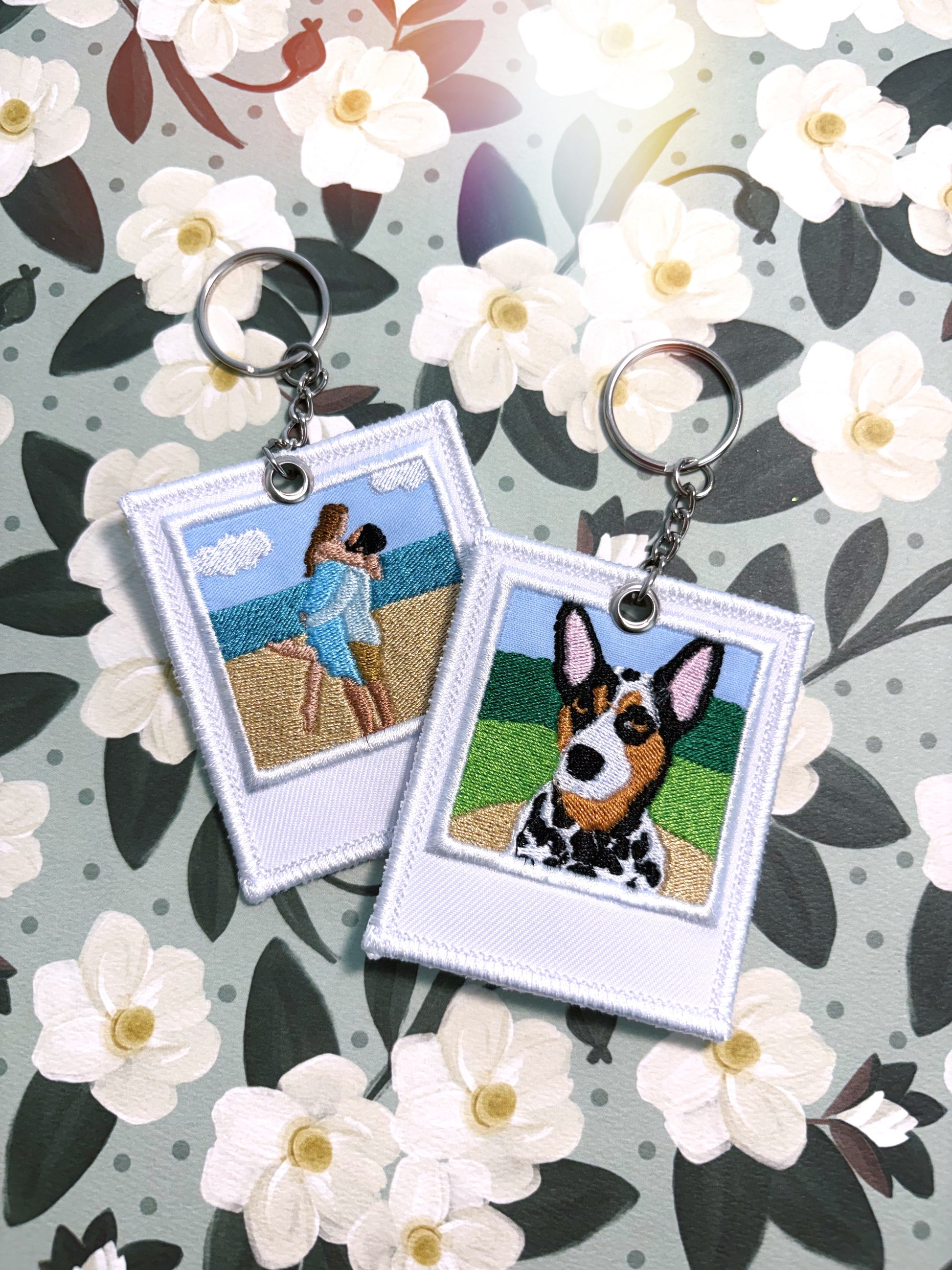 Two embroidered square keychains with silver key rings, displayed on a floral background. One keychain features an embroidered beach scene with two people, and the other features a black-and-white dog portrait.