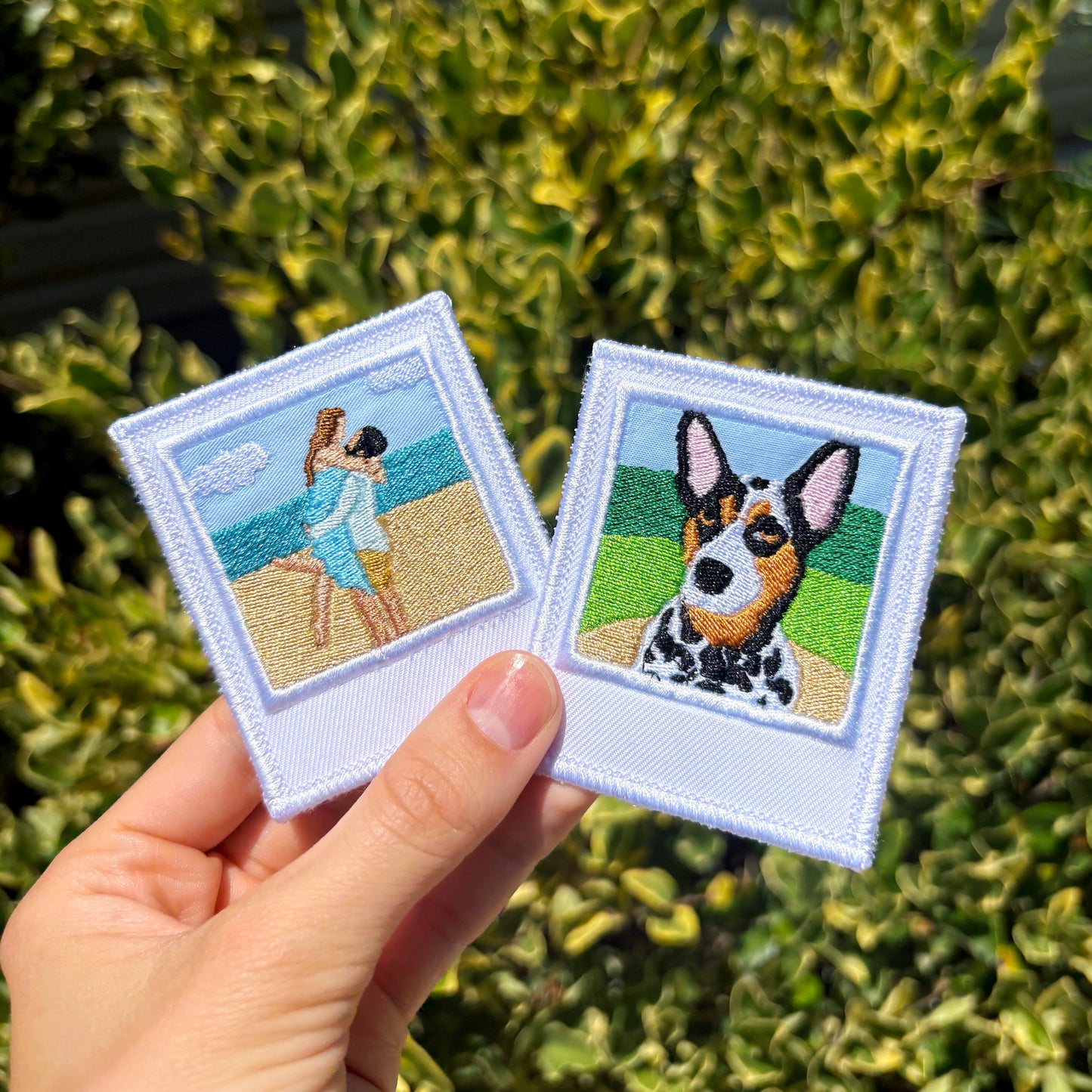 A hand holds two custom embroidered patches with white borders. One patch shows a couple on a beach, and the other shows a black-and-white dog with tan patches.