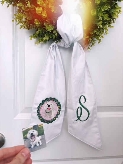 Wreath on a door that has a white wreath sash tied on it. Sash has embroidery of a dog on one side and “S” on the other.  