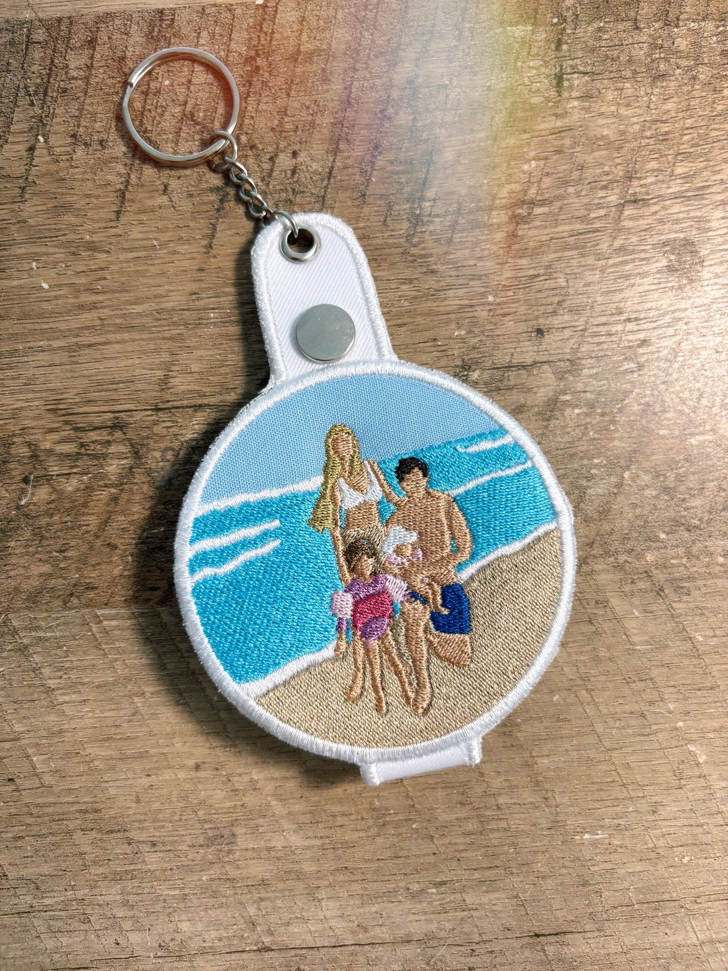 Keychain with embroidered design of a family on a beach, placed on a wooden surface.