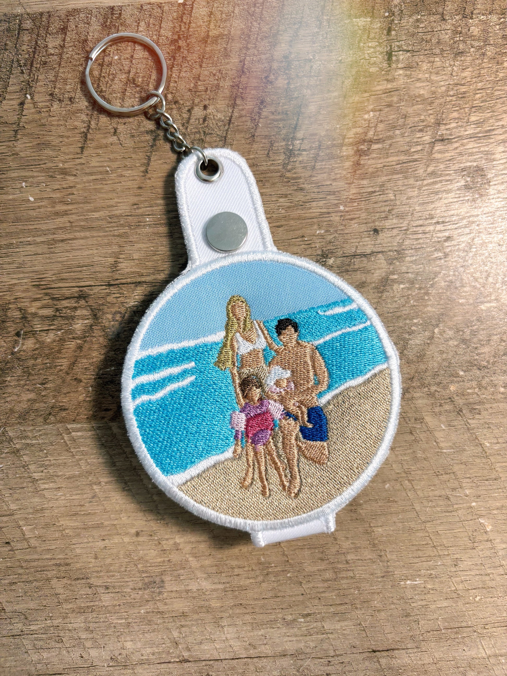 Keychain with embroidered design of a family on a beach, placed on a wooden surface.