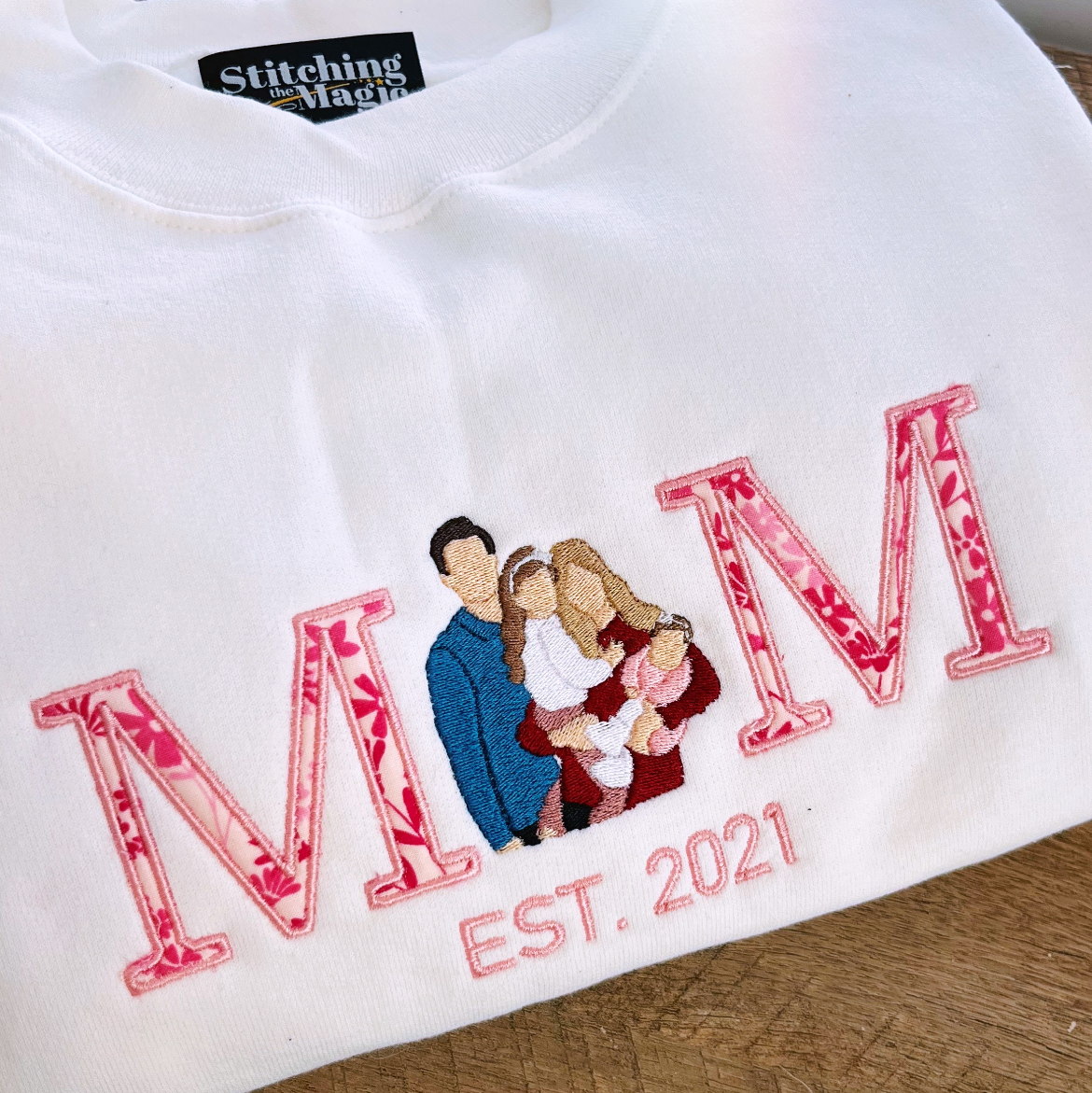 White shirt with 'MOM EST. 2021' text and embroidered family design on a wooden surface.