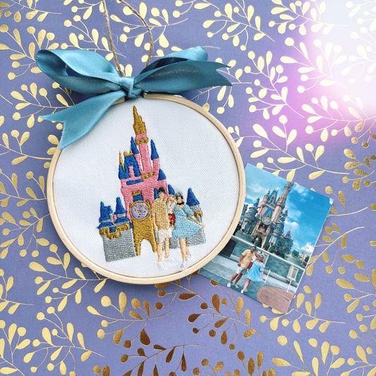 Ornament laying on a purple shiny background. Ornament features embroidery of a castle and a family. 