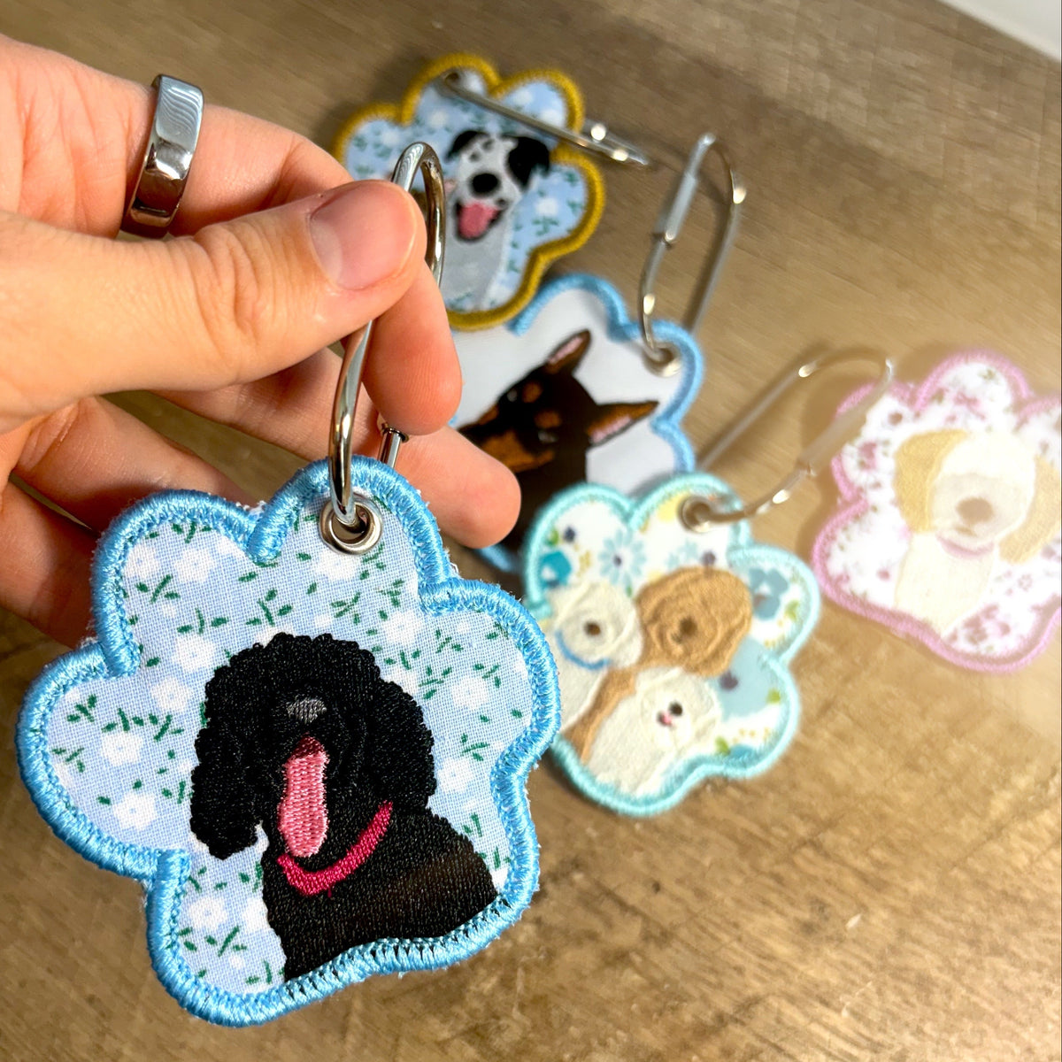 Wood table background with multiple keychains that are shaped like paw prints and have dogs embroidered on them.